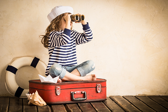 Kind in maritimer Kleidung sitzt auf einem Koffer und sieht durch ein Fernglas, daneben ein Rettungsring und eine große MuschelWei