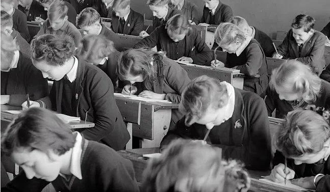 Bild einer alten Schulklasse mit Holzbänken, alle mit gesenktem Kopf über ein Blatt gebeugt. 