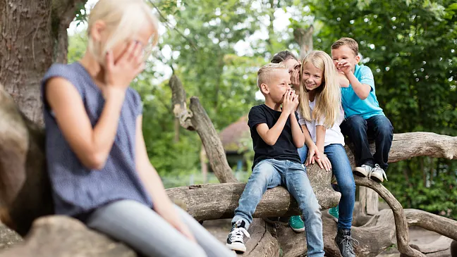 Kinder tuscheln über ein abseits sitzendes Mädchen, das sich die Hände vor das Gesicht hält