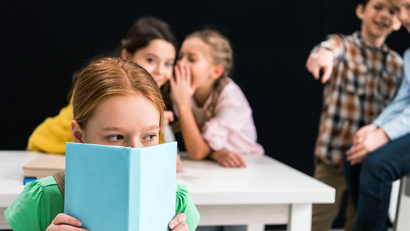 Kinder tuscheln hinter einem Mädchen, das sich hinter einem Buch versteckt