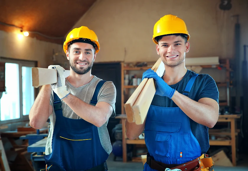 zwei junge Männer mit gelben Schutzhelmen und Blaumännern tragen zugeschnittene Holzlatten