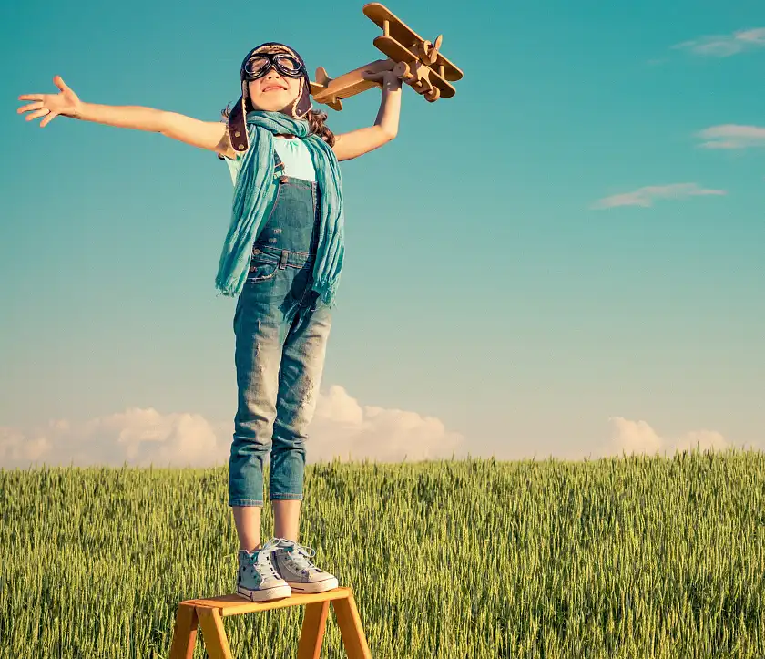 Ein Kind mit einer Pilotenmütze steht auf einer Leiter auf einer Wiese und hält ein Spielzeugflugzeug in der Hand